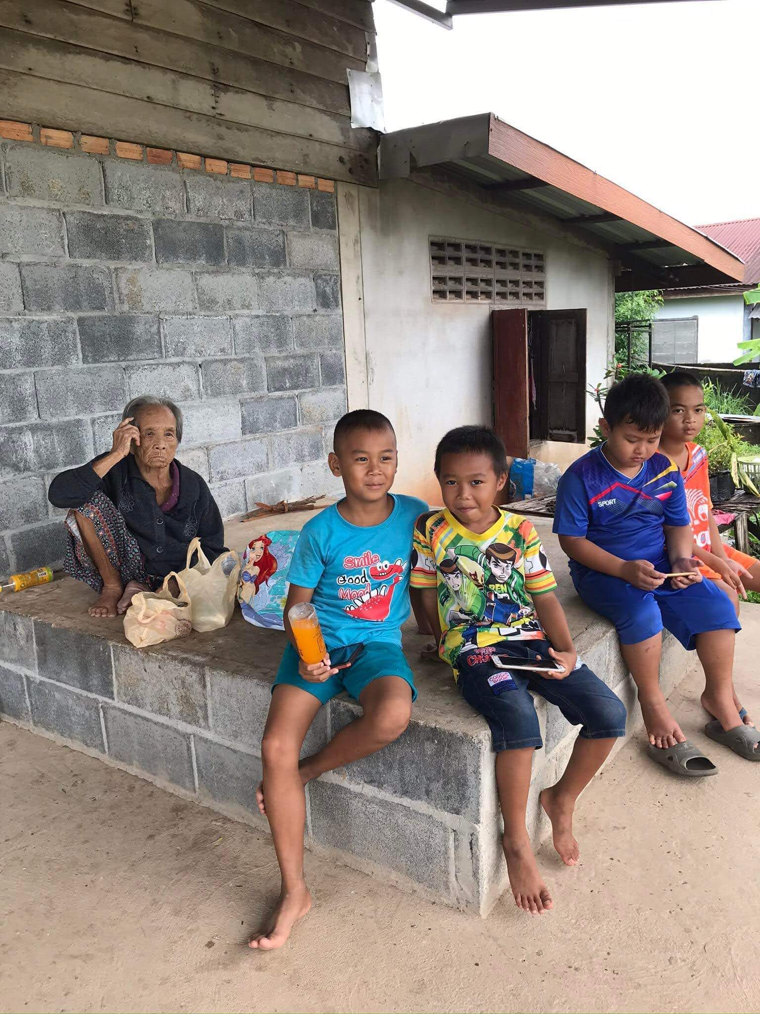 a Thai elder woman sits on her front porch behind a young boy and his friends