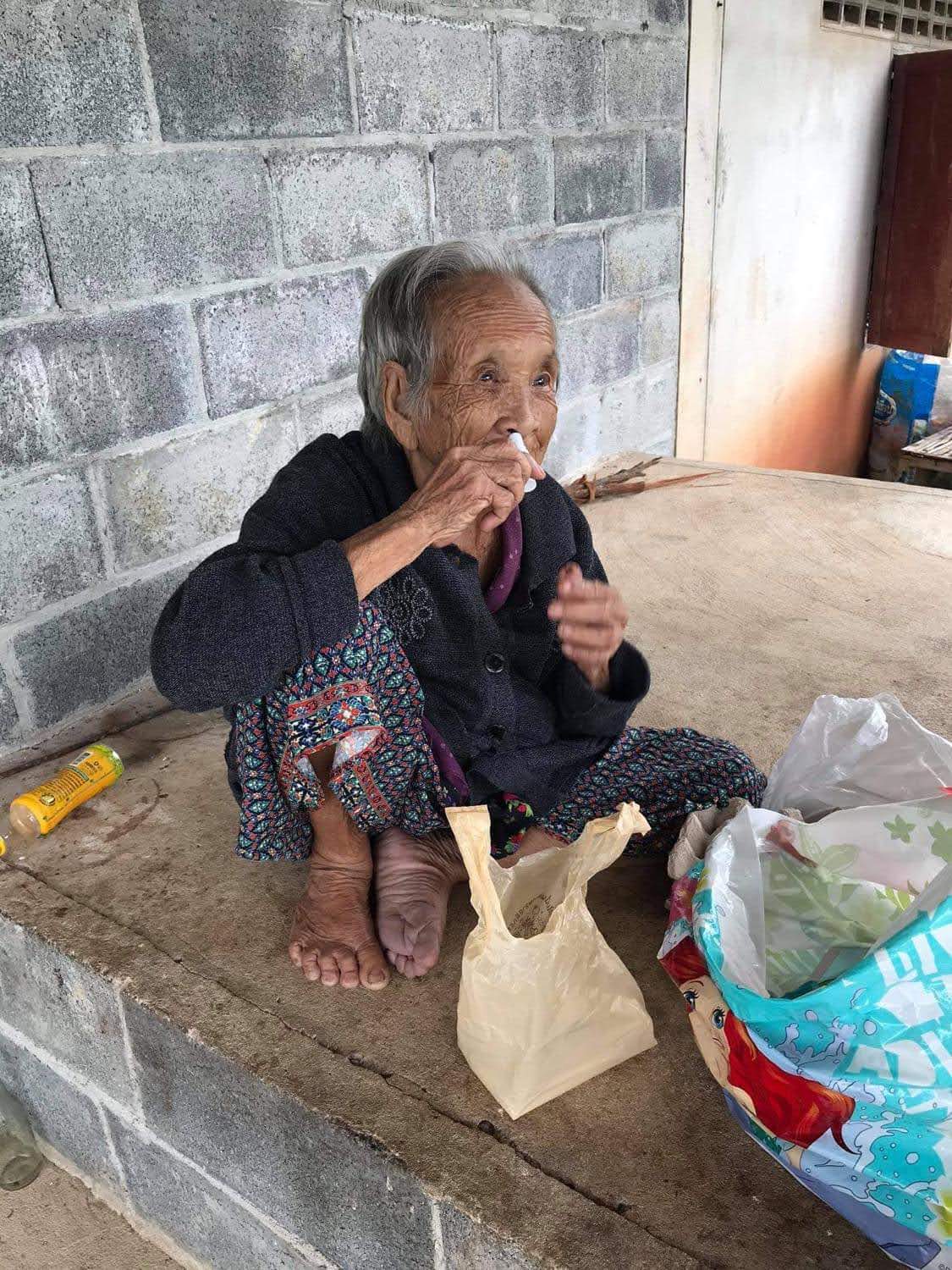 a Thai elder woman sits with a bag of food in front of her and sniffing an inhaler