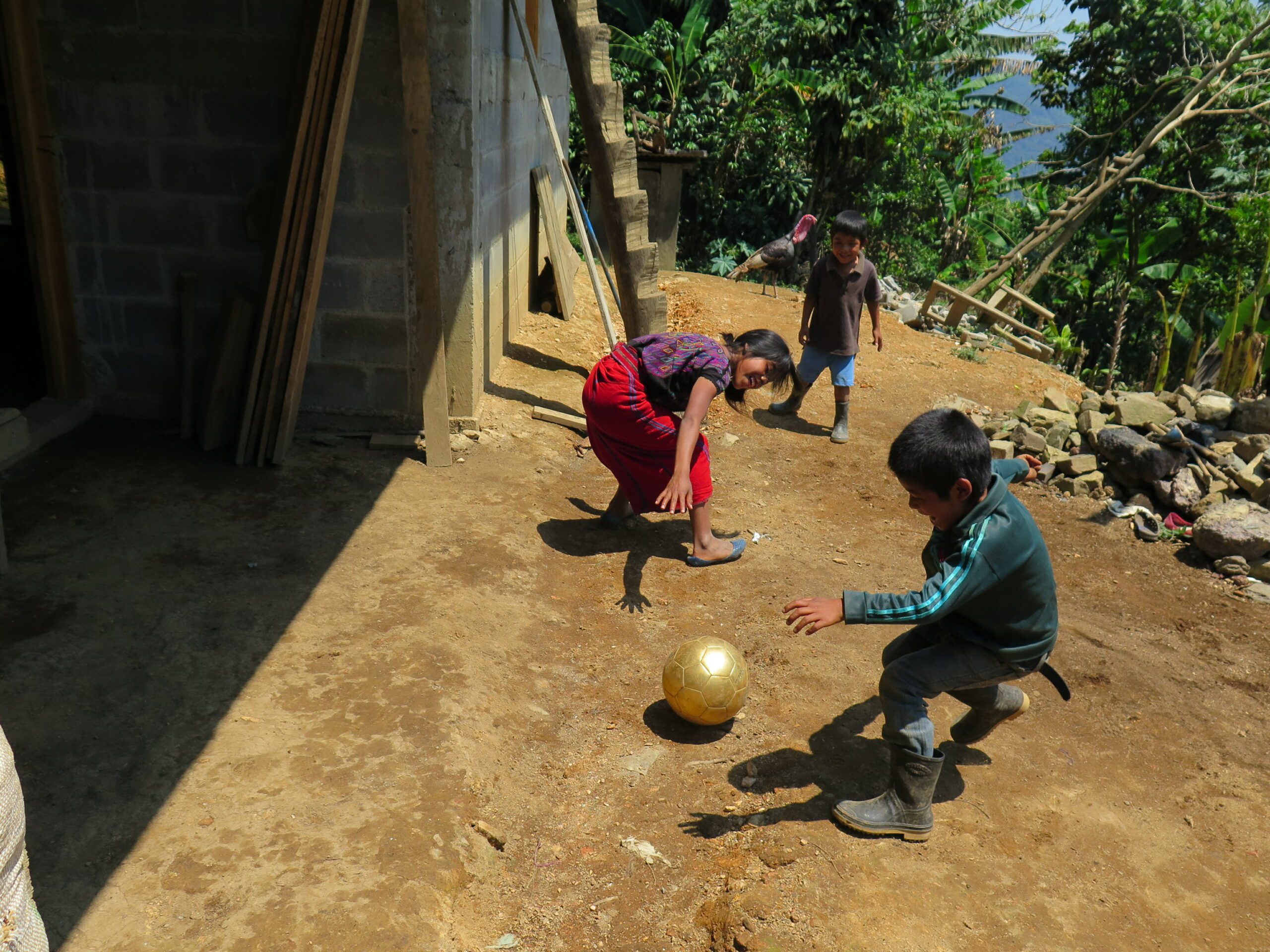 a couple of young kids kick a ball in the dirt in front of their house