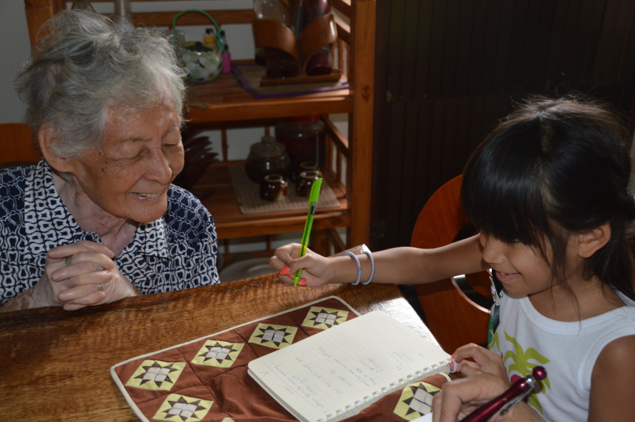 an elderly woman, Vanida, sits with a young Thai girl at a desk looking at something together